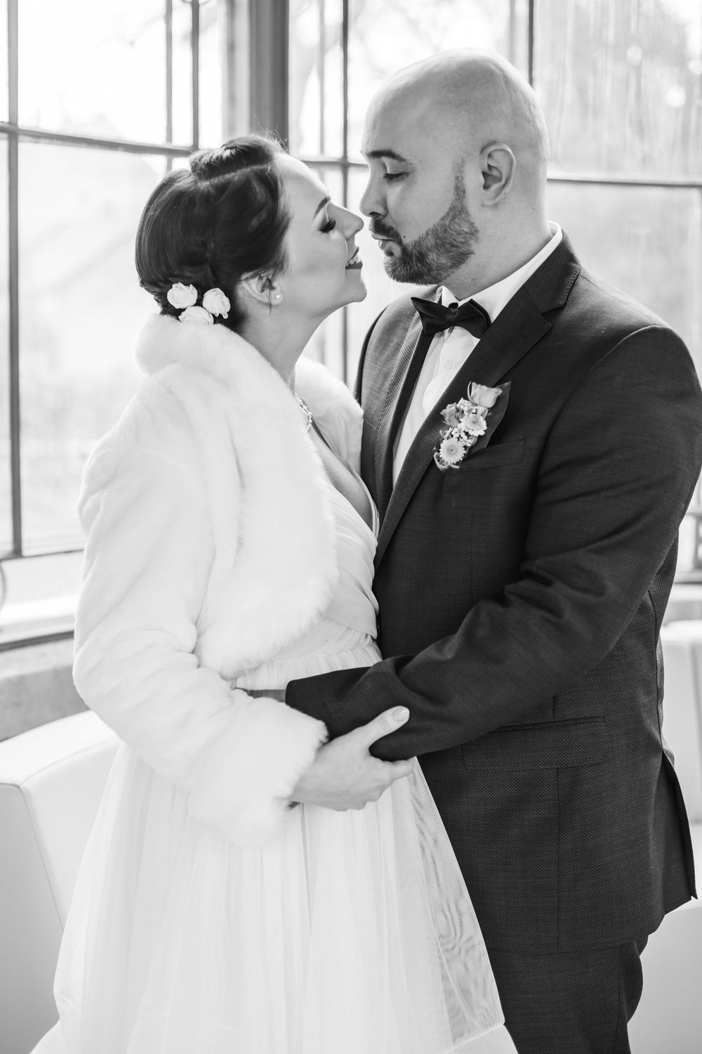 Black and white wedding portrait of couple in formal attire sharing intimate moment near window.