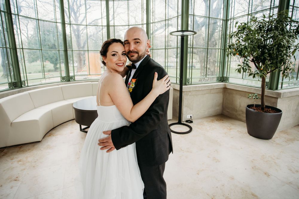 A couple in wedding attire share a romantic embrace in a bright sunlit conservatory with large windows and potted plants.