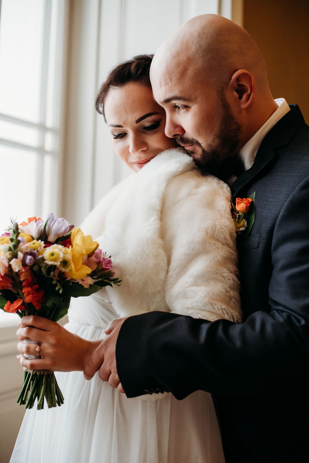 Newlywed couple embraces by window with colorful autumn bouquet, bride wearing white dress with fur wrap.