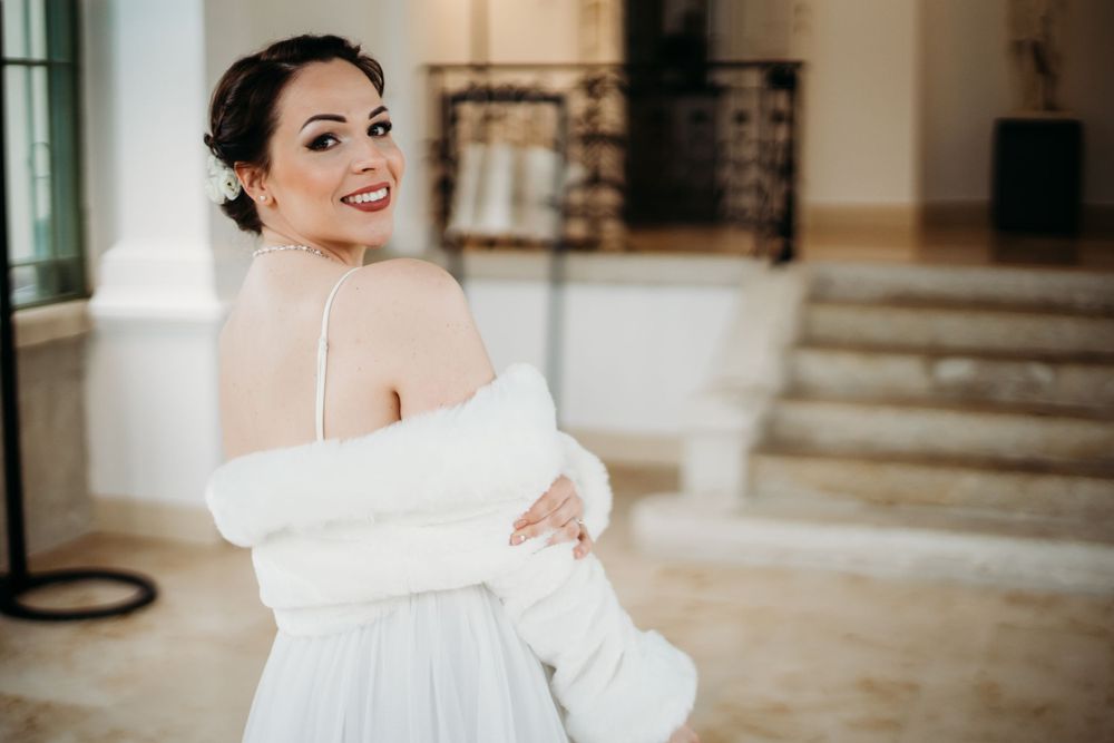 A bride wearing a white strapless gown and cozy cardigan wrap smiles warmly in an elegant interior setting.
