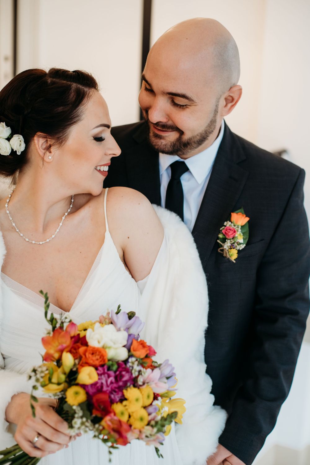 A bride in white holding a vibrant multicolored bouquet shares an intimate moment with a groom in a dark suit and tie.