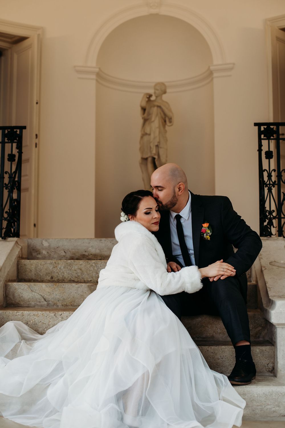 Elegantly dressed couple in wedding attire sitting together on stone steps with classical statue in alcove behind them.