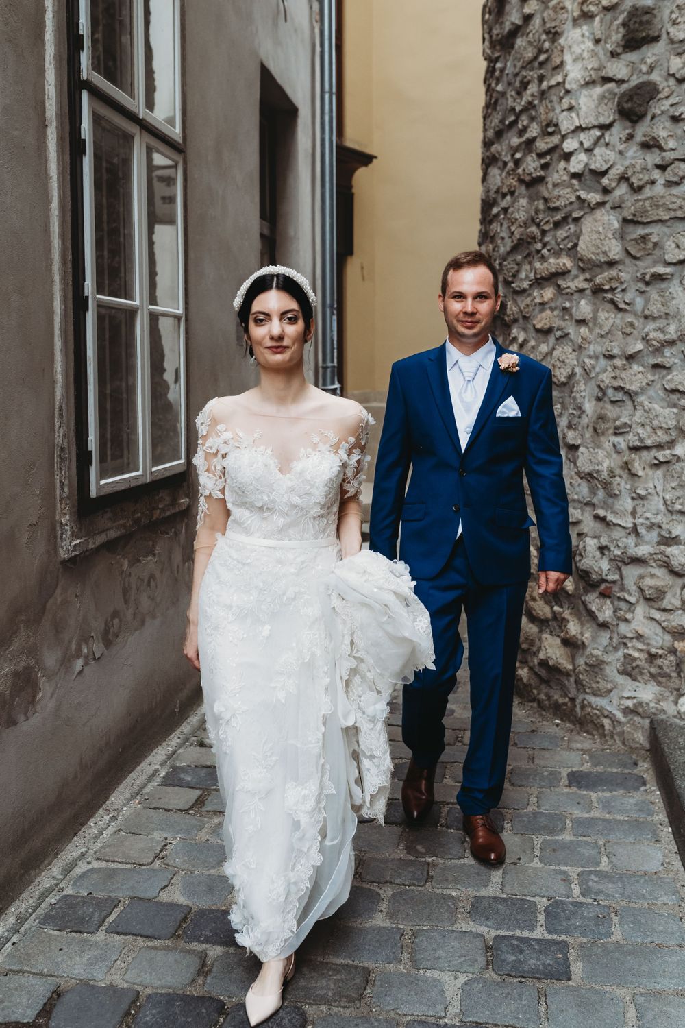 A couple in wedding attire walk together on a cobblestone street beside an old stone wall.