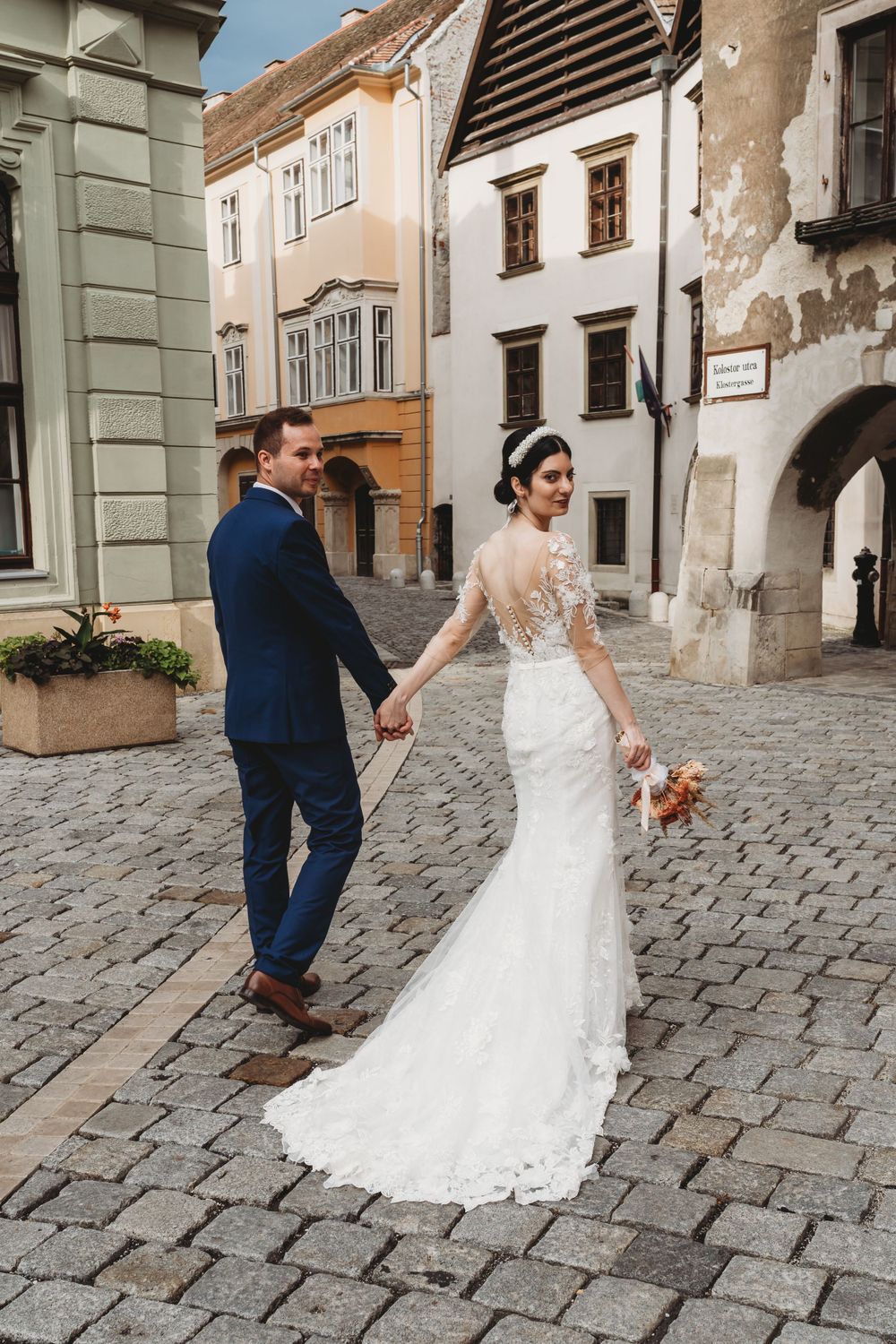 A newlywed couple walking on a cobblestone street in a historic European town with aged buildings in the background.