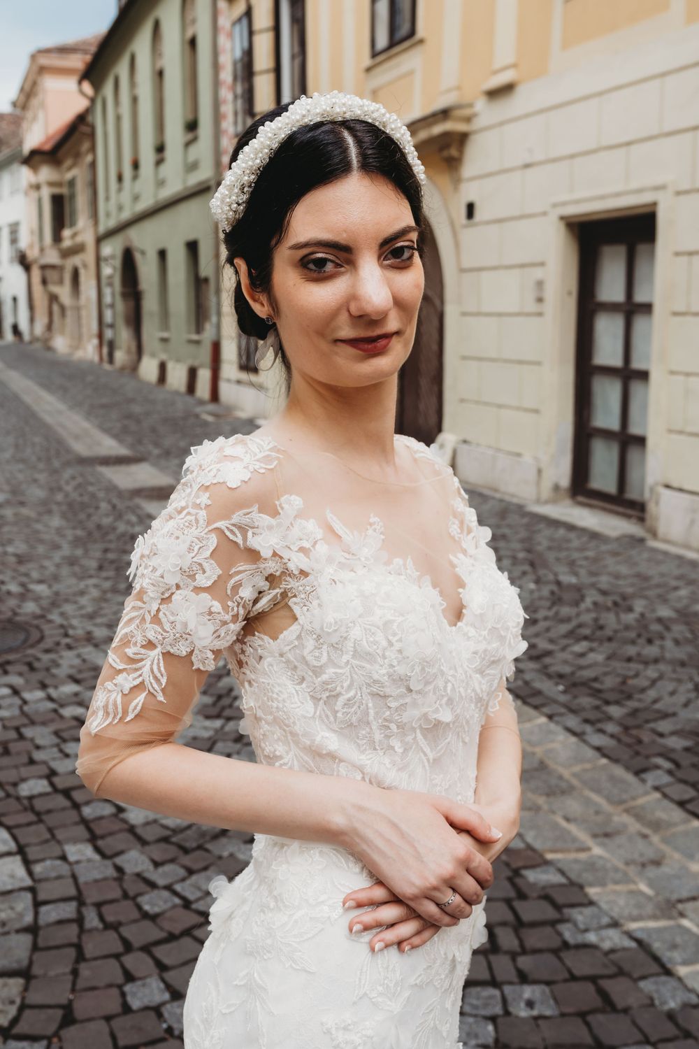 Elegant lace wedding dress with sheer sleeves photographed on a cobblestone street with historic buildings in background.