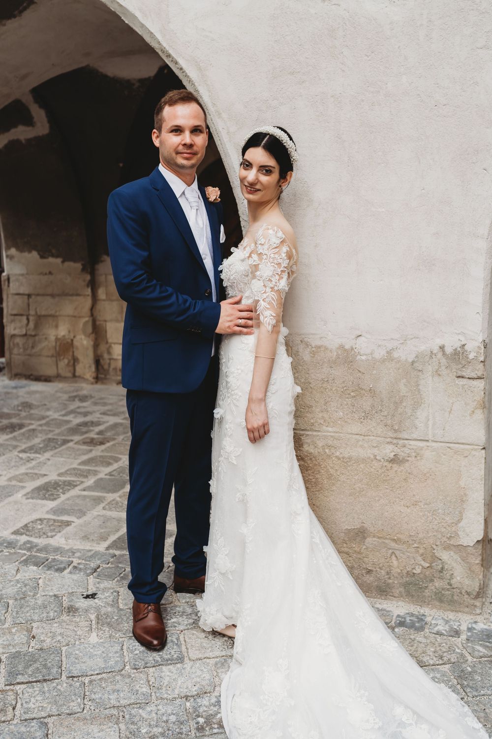 A bride and groom pose together against a stone wall, with the bride wearing a lace dress and the groom in a navy suit.