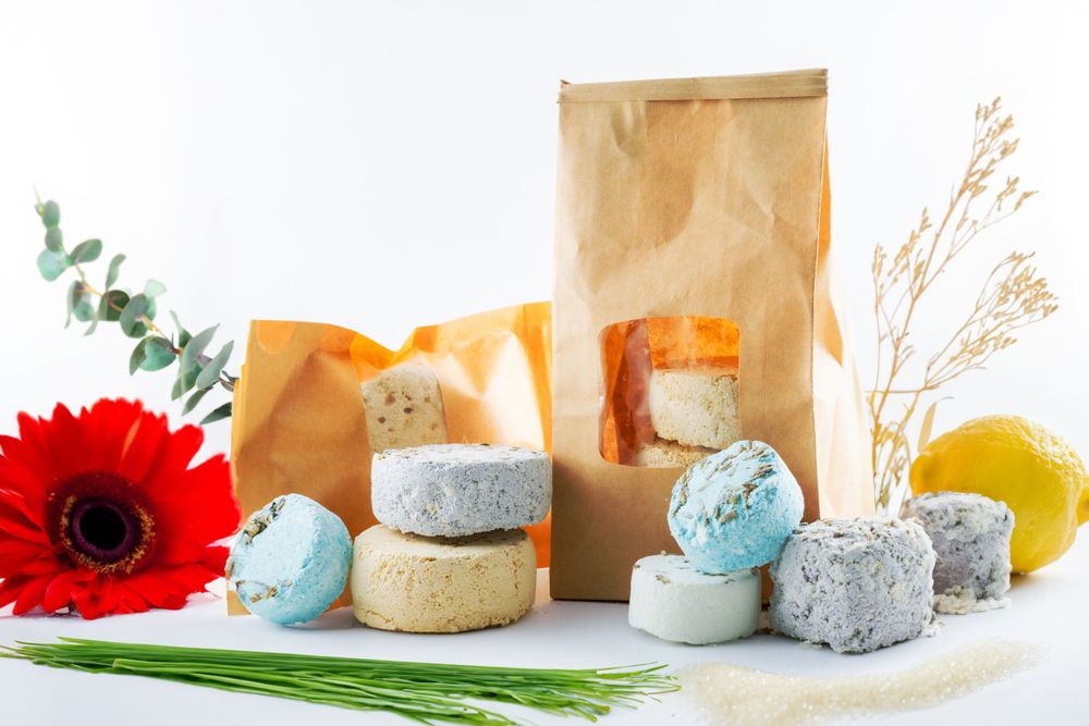 Round handmade bath bombs and soap bars displayed with a red gerbera daisy, brown paper bags, and floral decorations.