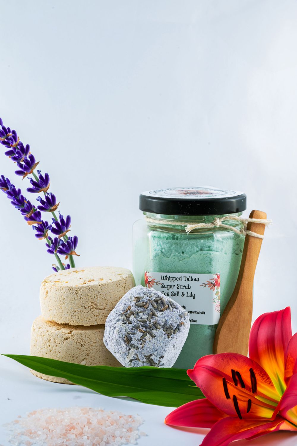 Spa beauty products with lavender flowers, sea salt scrub jar, natural bath bombs, and vibrant lily flower on white background.