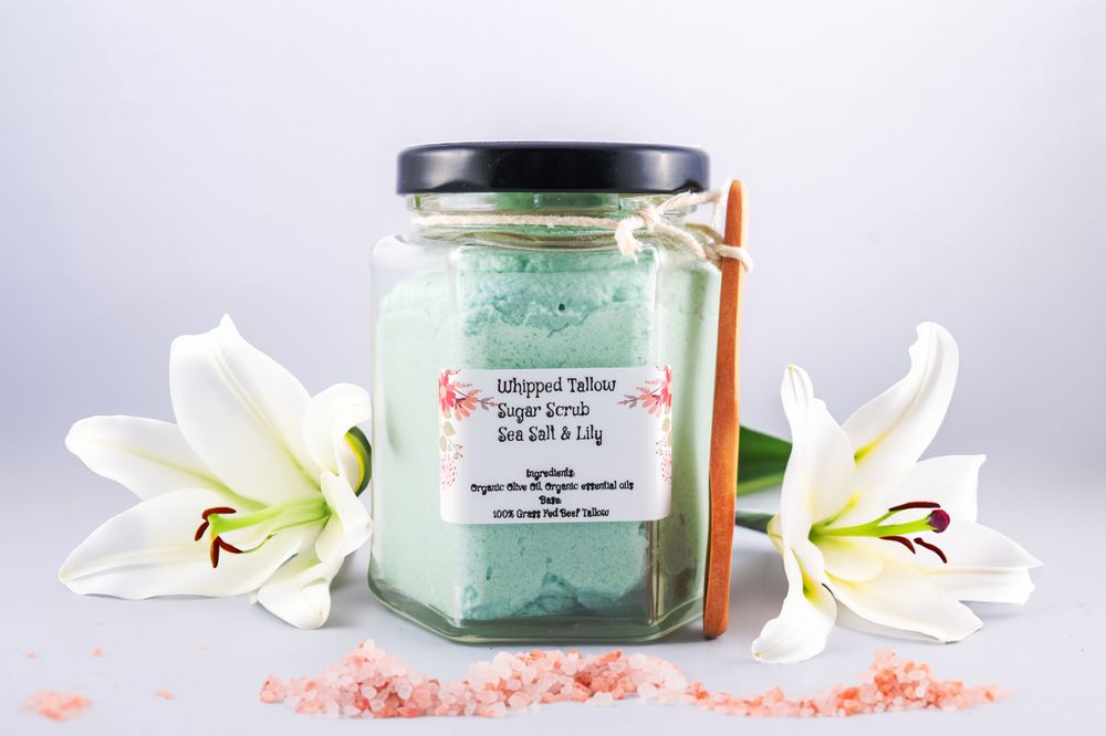 Spa jar with body scrub surrounded by white lilies and pink Himalayan salt on a light gray background.