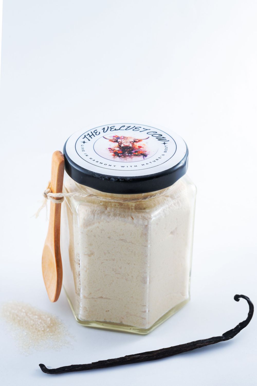 Glass jar filled with homemade vanilla sugar alongside a wooden spoon and dried vanilla bean on white background.