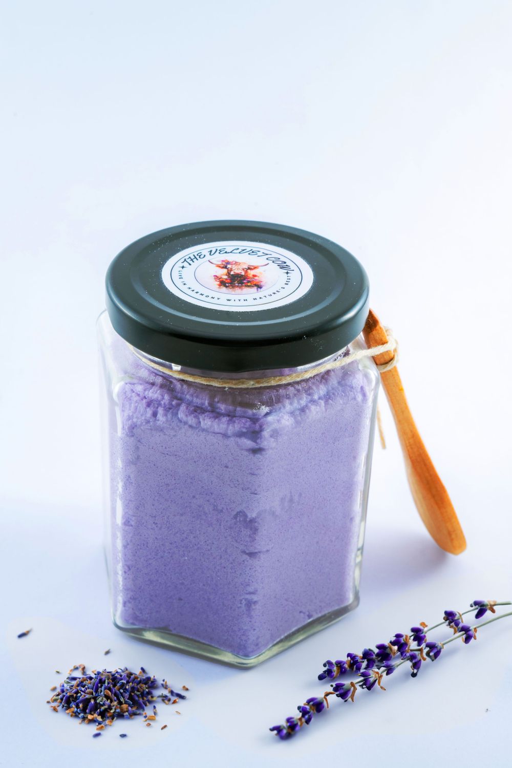Glass jar filled with purple lavender bath salt next to dried lavender sprigs on light background.