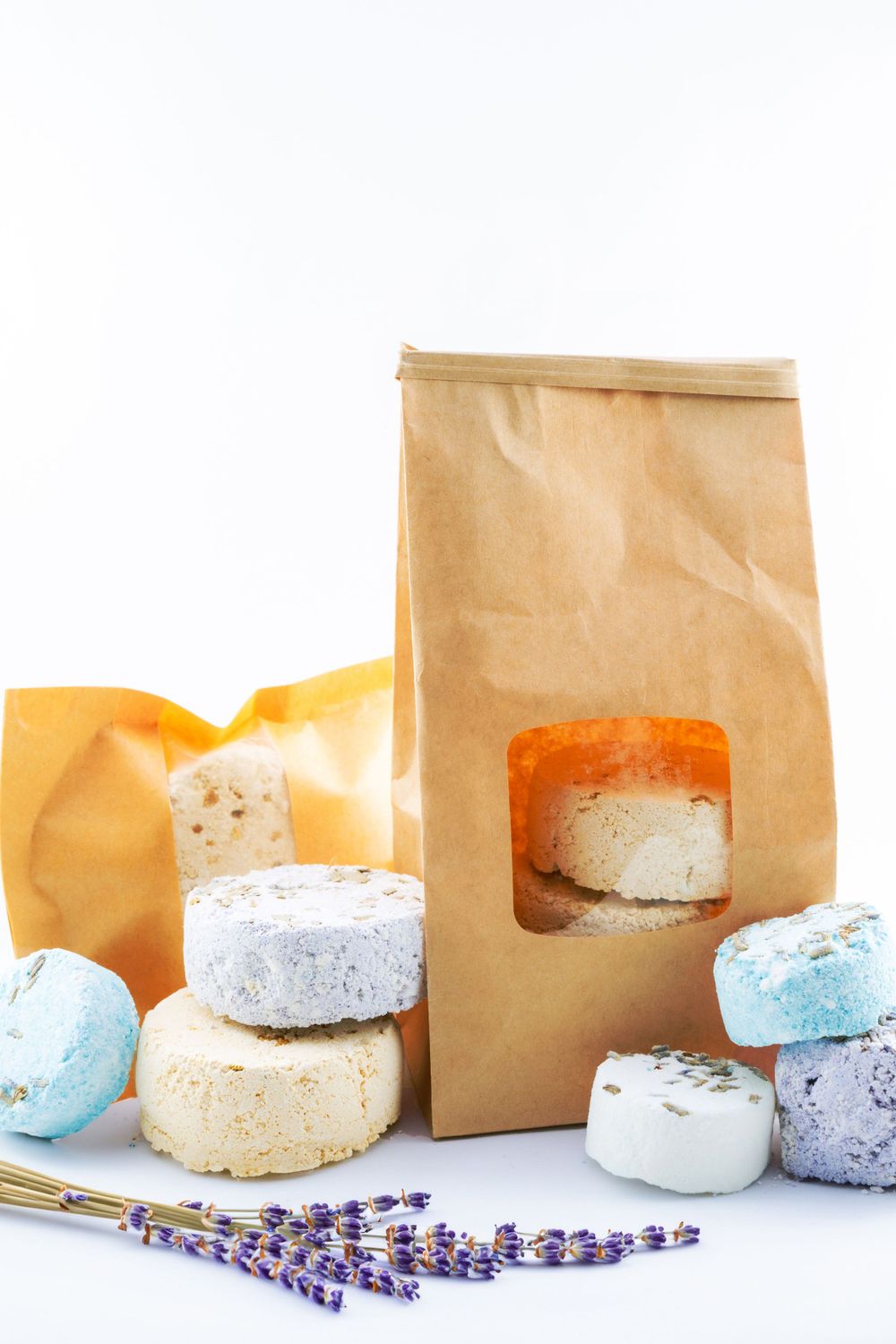 Bath bombs and soaps in various colors with lavender stems and brown paper bag against white background.