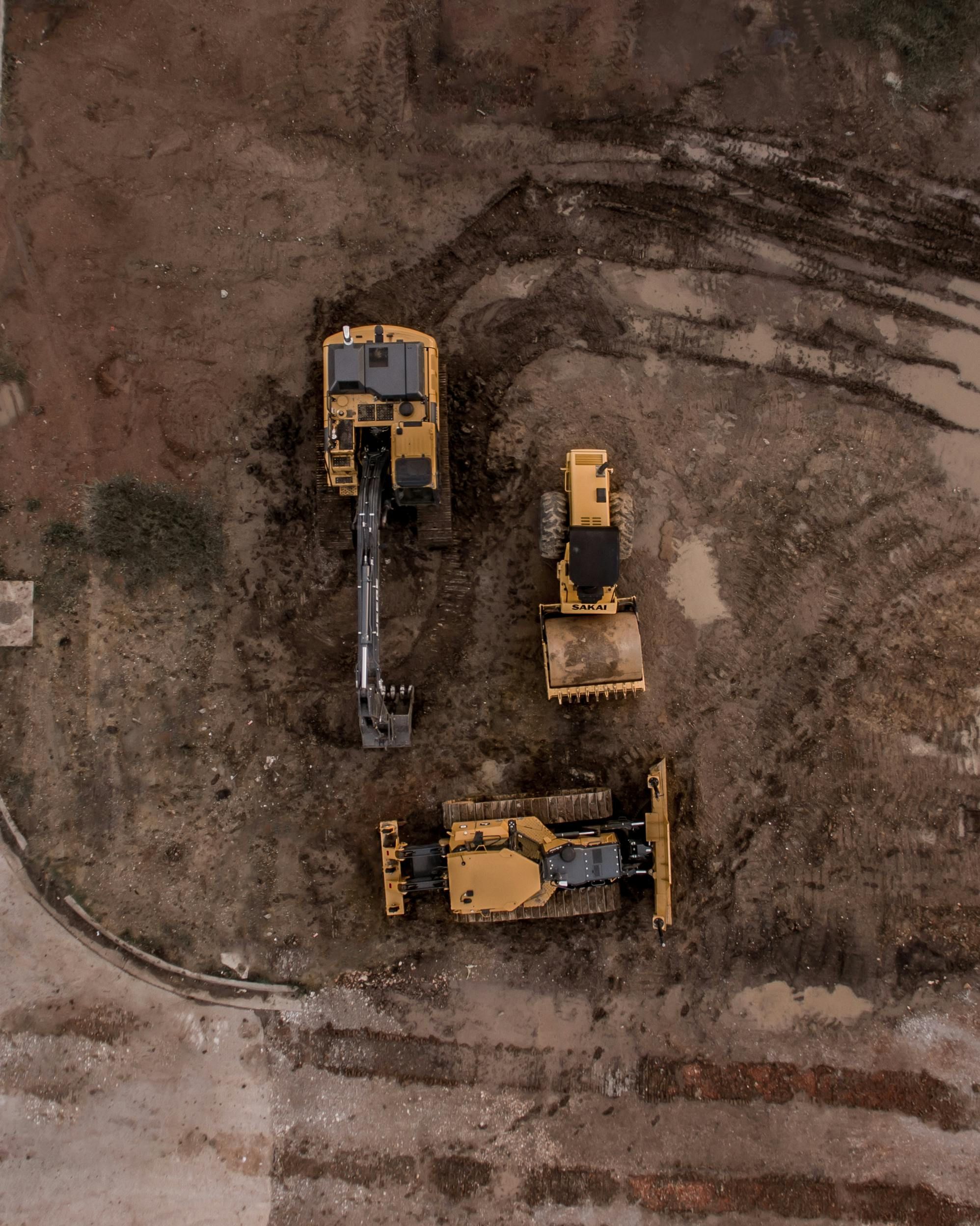 Aerial view of three yellow construction vehicles parked on brown dirt ground at a worksite.