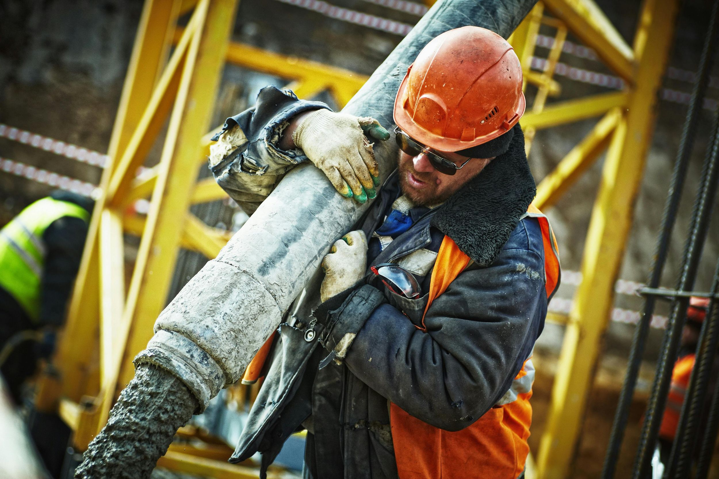 Worker in orange safety vest and hardhat carrying heavy industrial pipe at construction site.