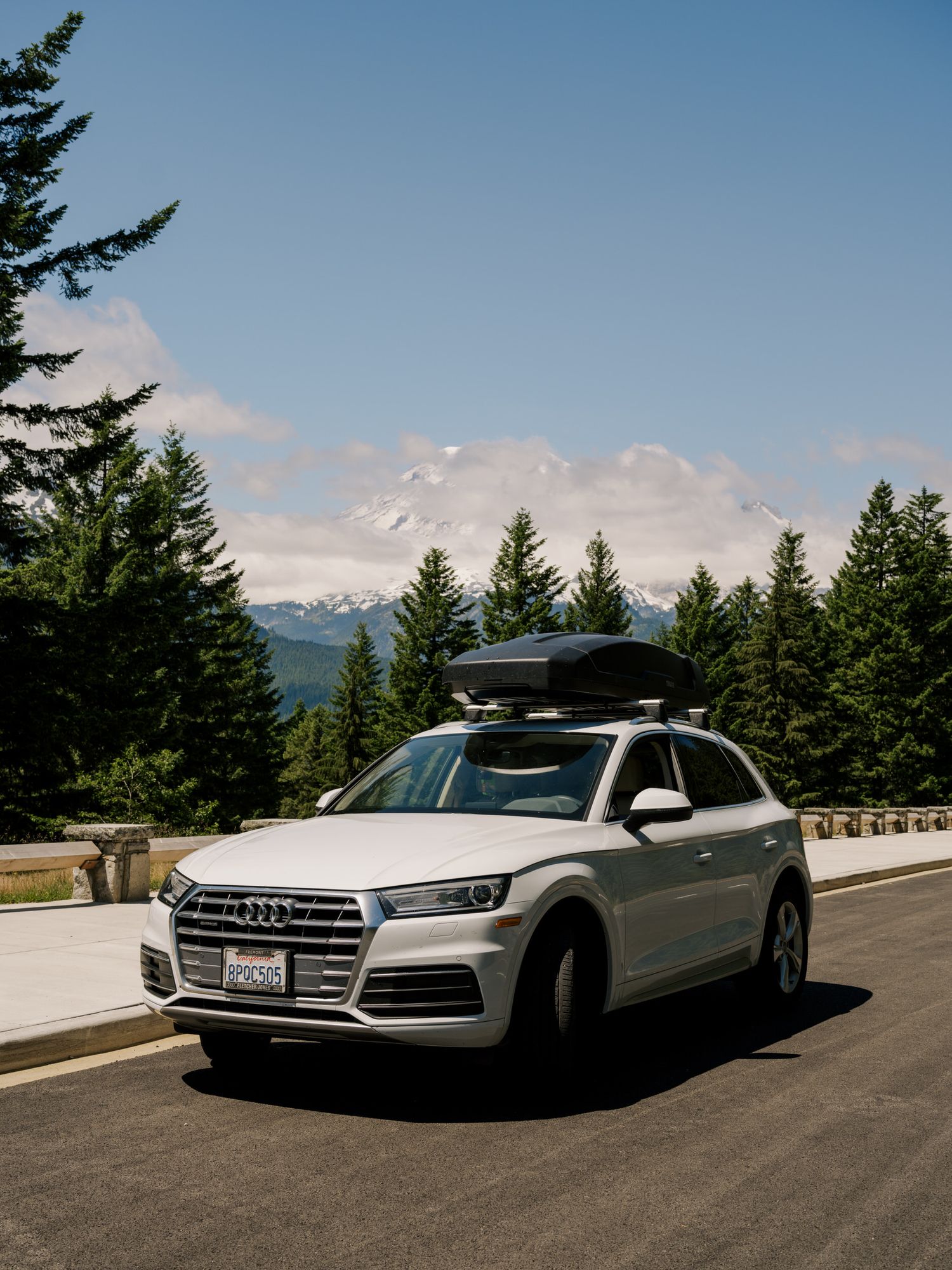 White Audi SUV with roof cargo box drives along mountain road surrounded by evergreen trees and scenic views.