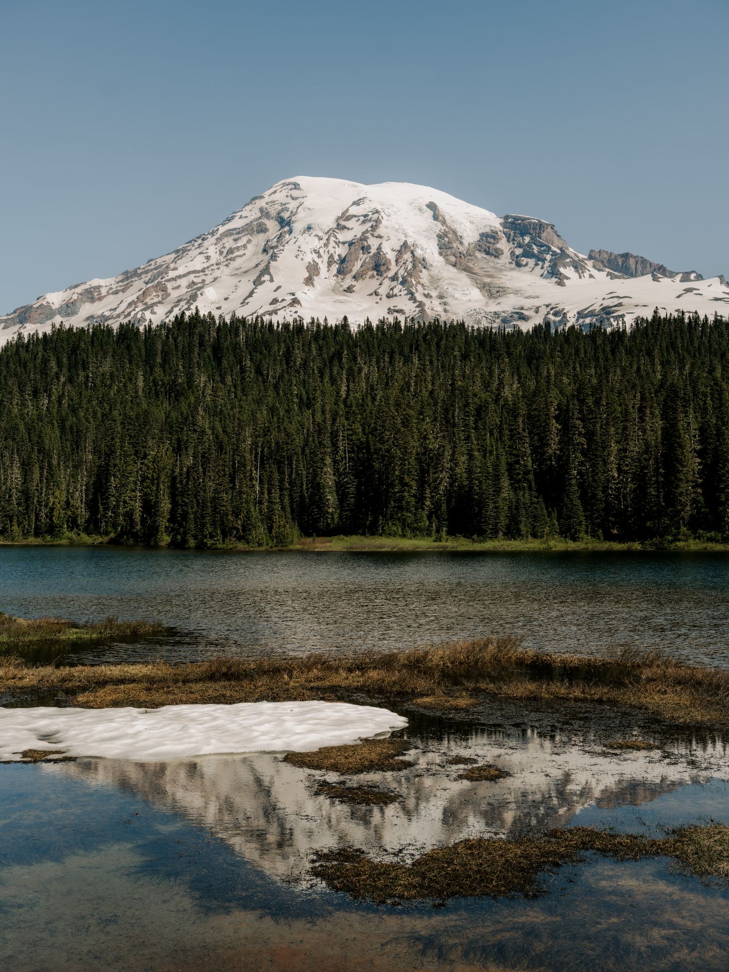 Snow-capped Mount Rainier reflects in a pristine alpine lake surrounded by dense evergreen forest under blue sky.