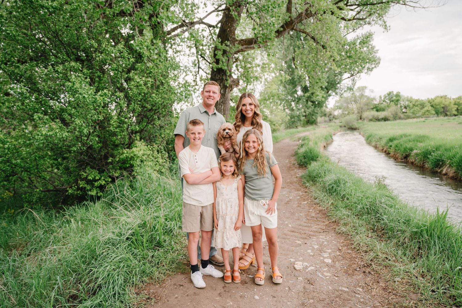 Group stands together on a dirt path alongside a stream with green trees and vegetation in the background on a sunny day.