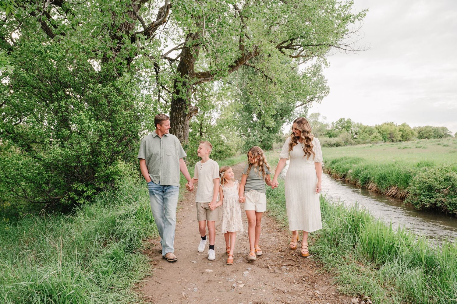 A family holds hands while walking along a peaceful dirt path beside a stream surrounded by lush greenery on a summer day.