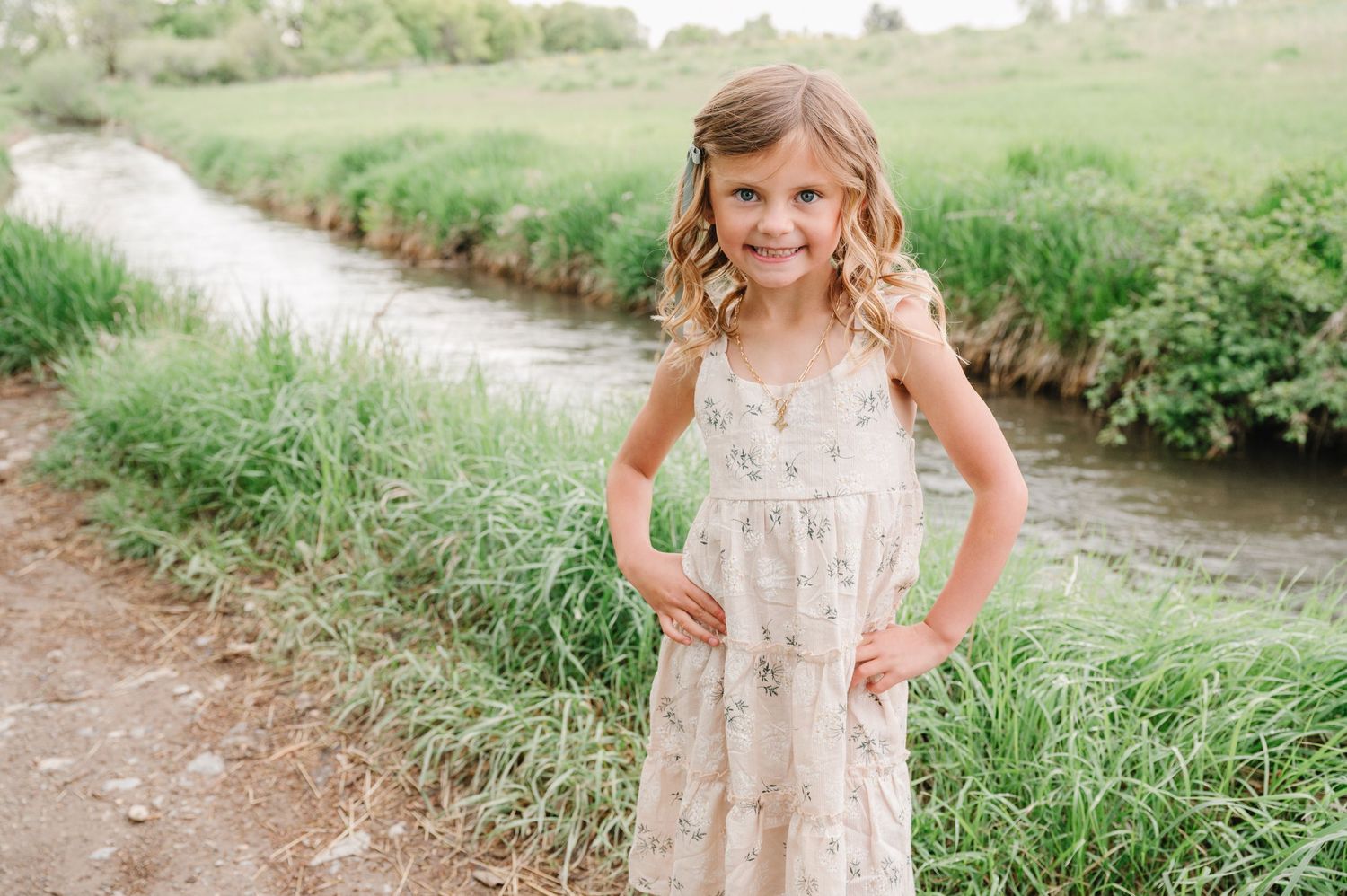 A child in a floral dress stands along a peaceful creek bank surrounded by tall green grass on a sunny day.