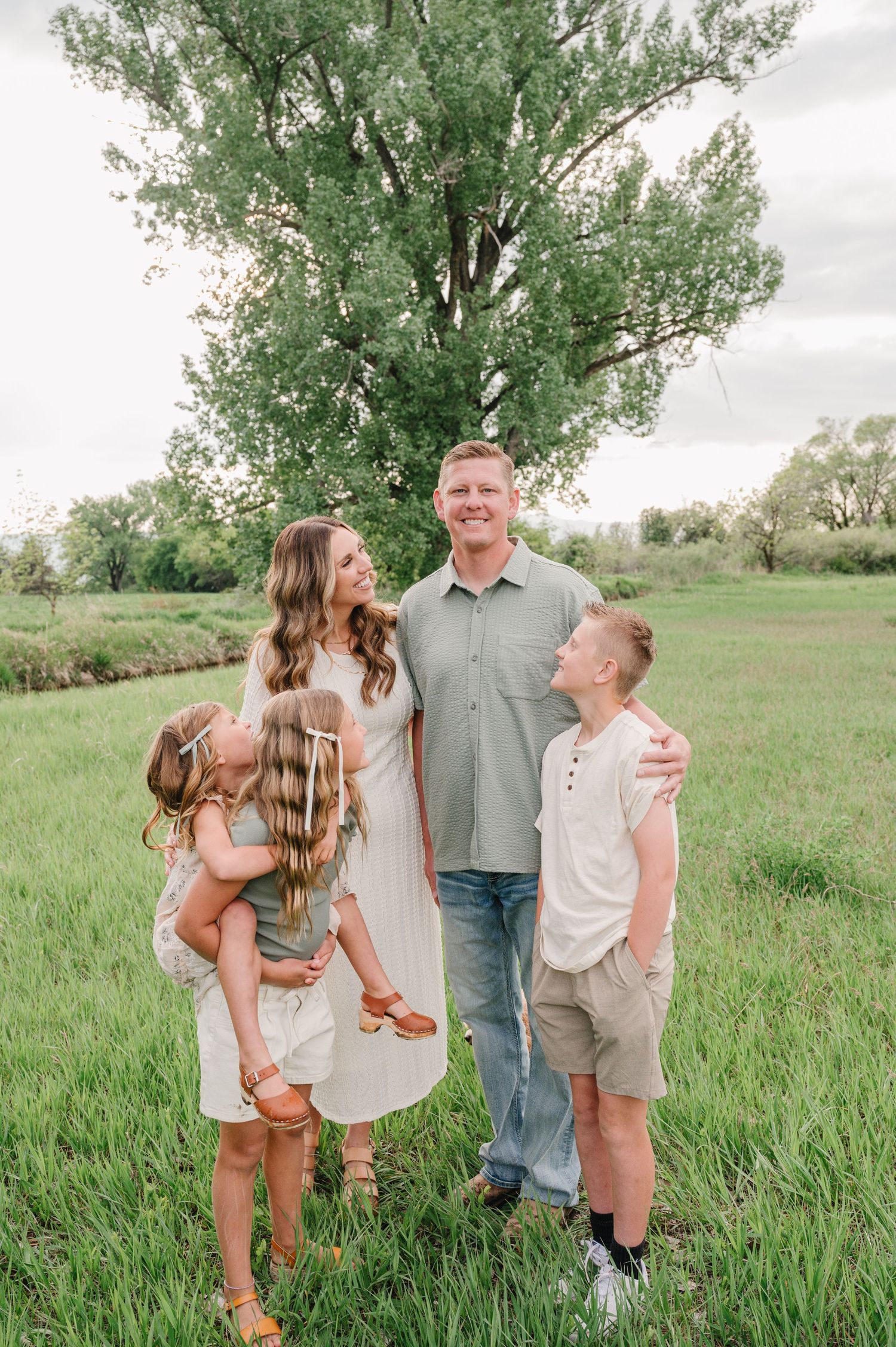Family group in casual clothing standing together in a lush green field with a tall tree in the background on a sunny day.
