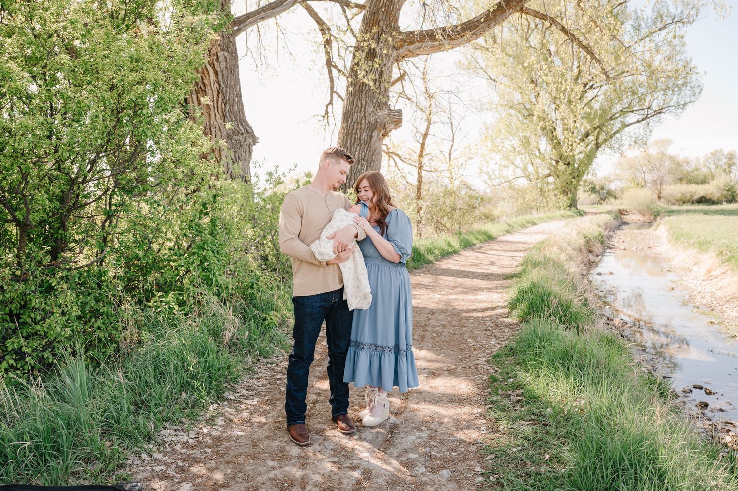 A couple stands on a scenic path by a stream, cradling their newborn baby surrounded by lush green trees.