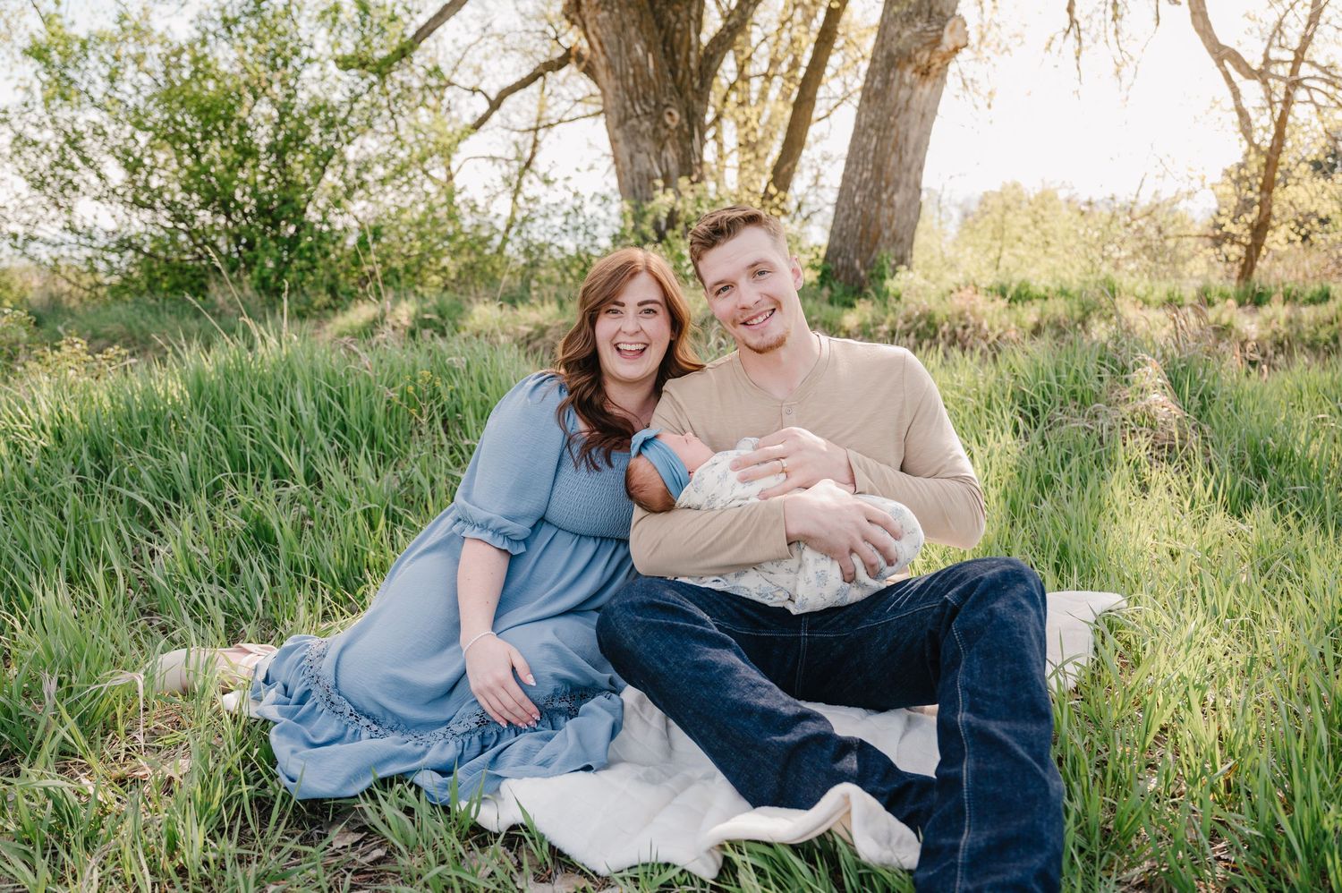 A young couple sits on a blanket in a green meadow, smiling while holding their newborn baby outdoors.