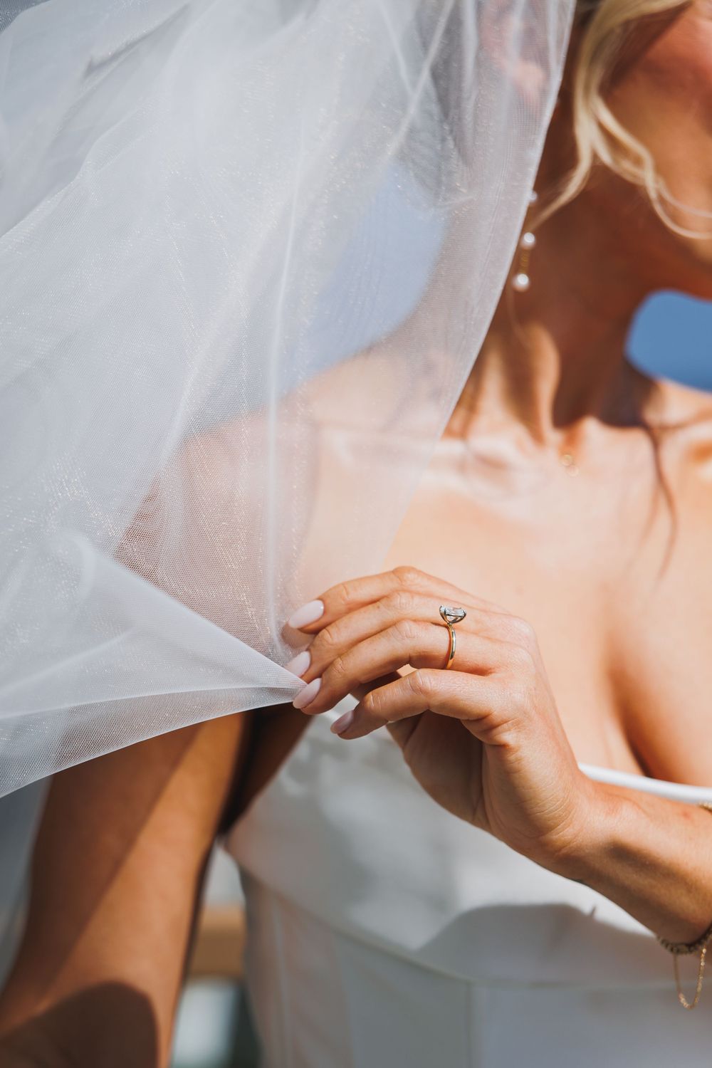 Bride in white wedding dress holding veil, wearing a diamond engagement ring, with soft natural lighting.