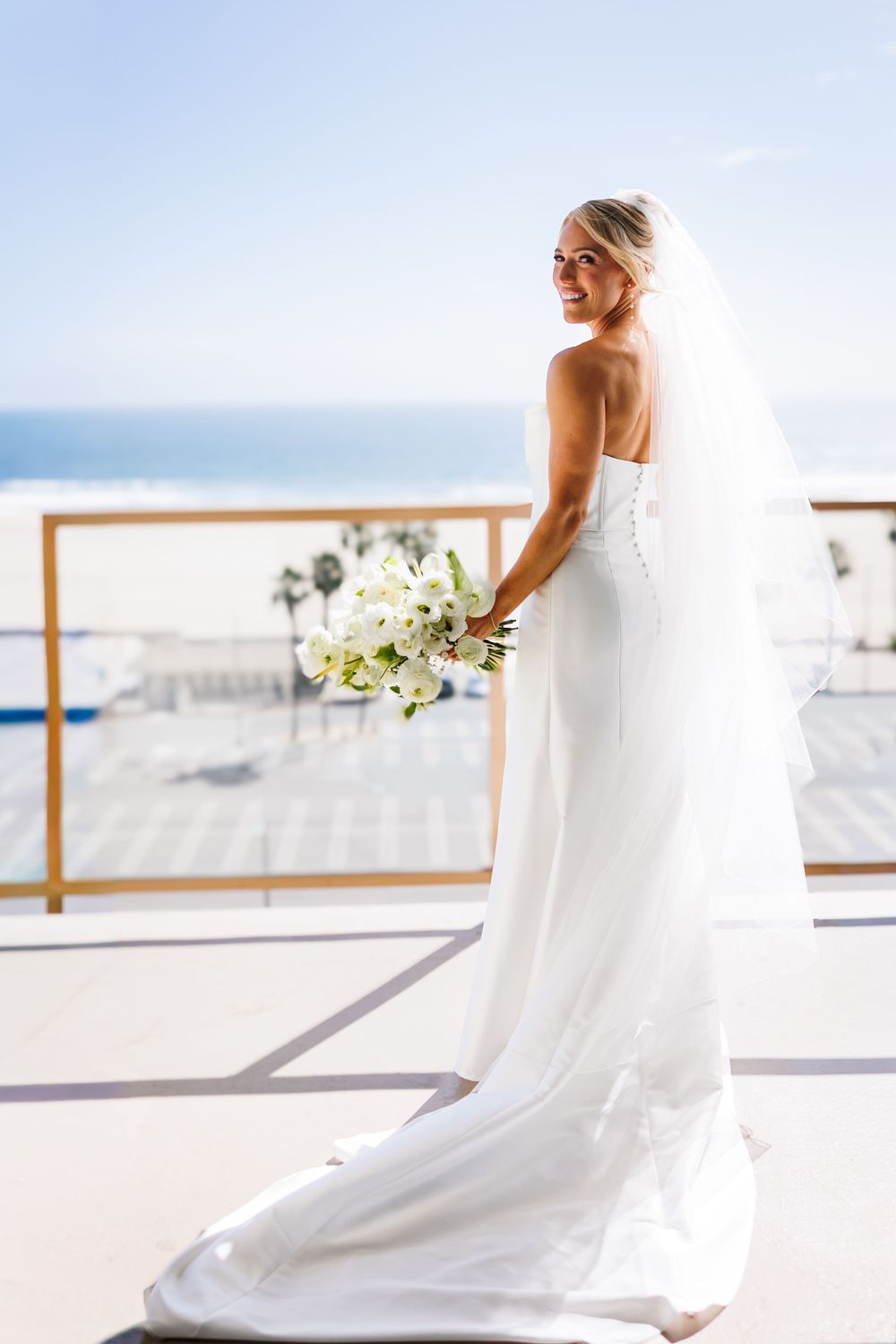 Smiling bride in strapless white gown and veil holds white bouquet on sunny oceanview terrace.