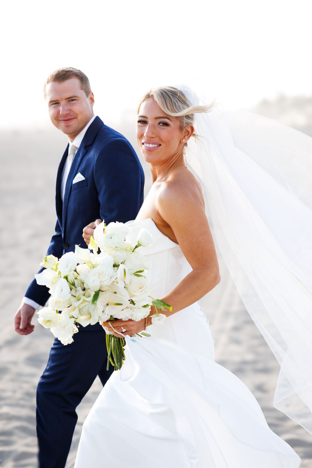 Smiling bride in white strapless gown and veil holds bouquet of white flowers while groom in navy suit walks behind on beach.