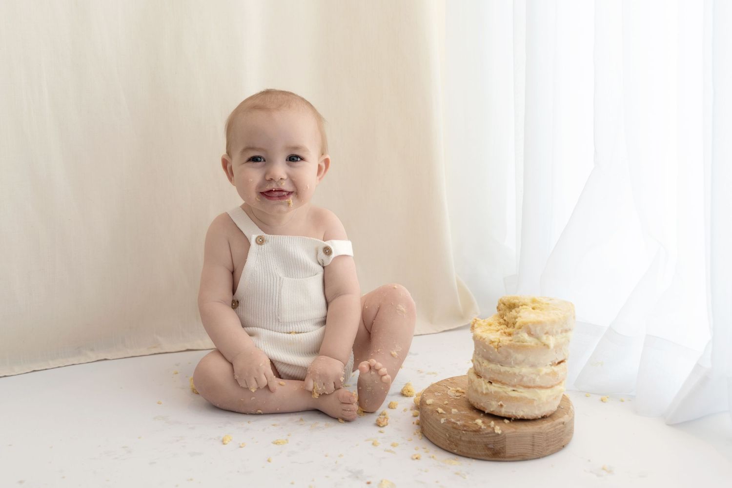 Baby in white romper sits next to a rustic naked cake during milestone photoshoot session against white backdrop.