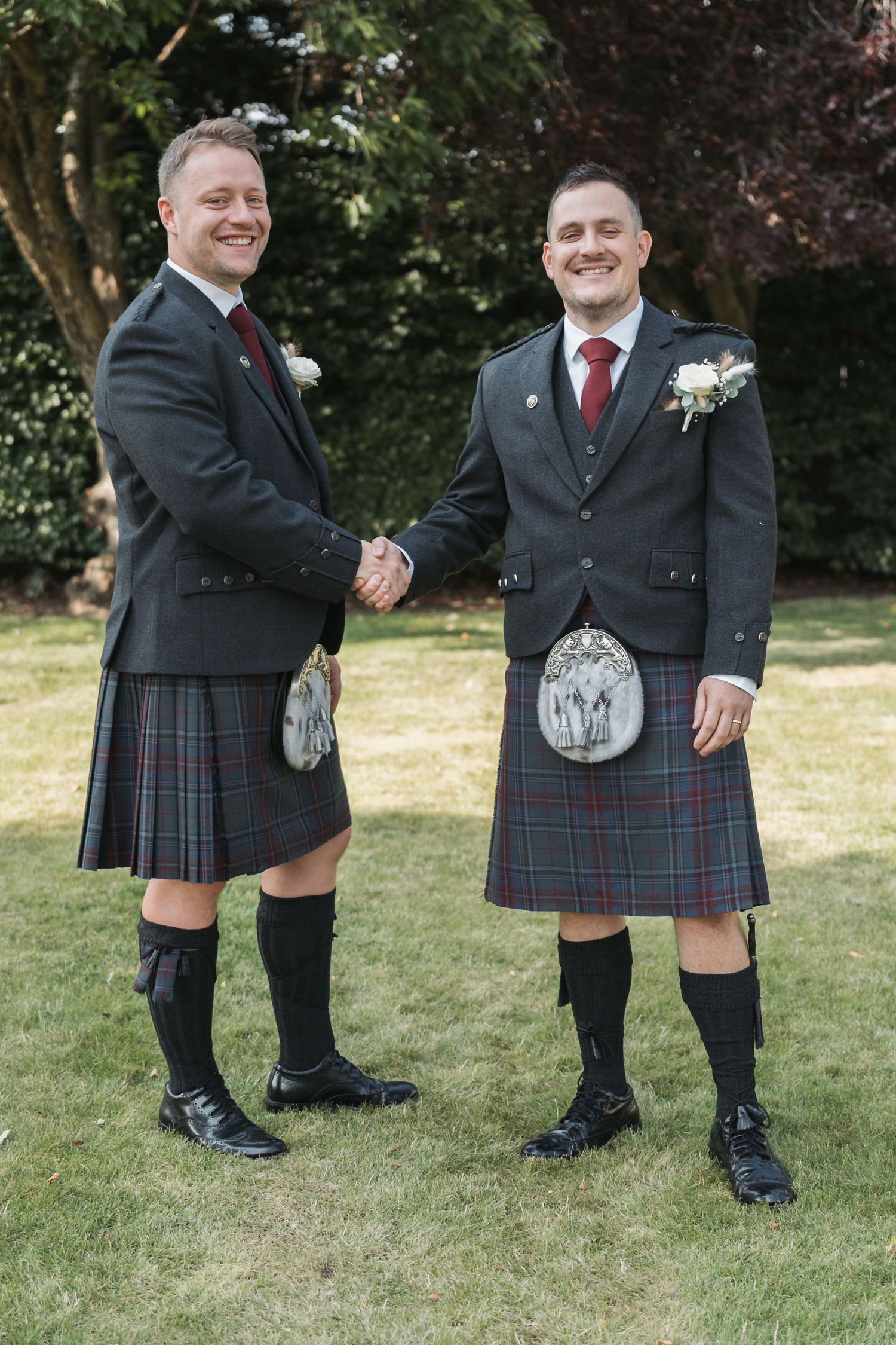 Two men in traditional Scottish kilts and formal jackets shake hands while standing on grass at an outdoor event.
