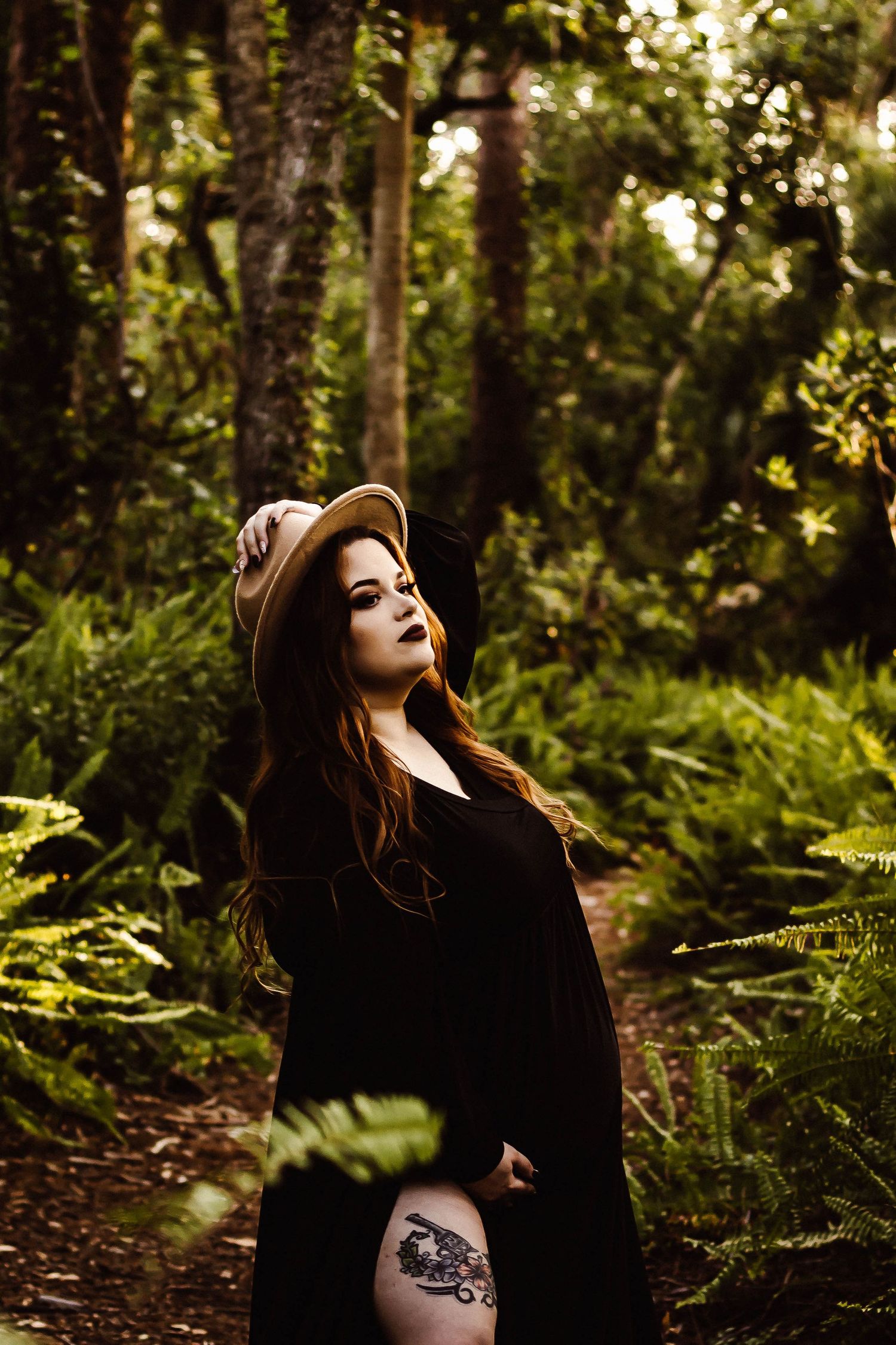 Woman in dark clothing poses dramatically in a sunlit forest with ferns.