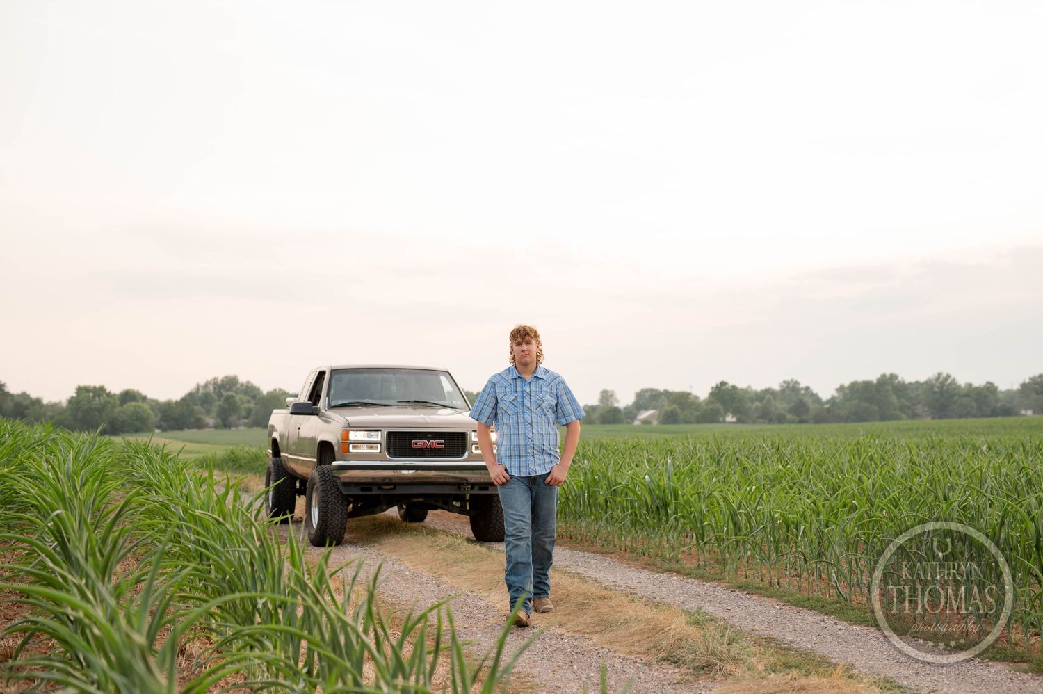 Person wearing plaid shirt walking on dirt road between cornfields with GMC truck behind.
