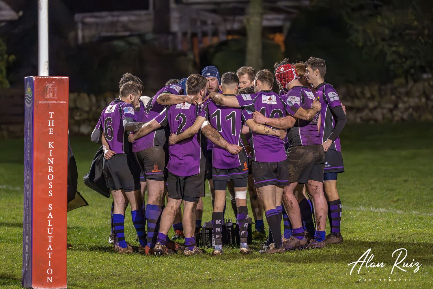 Rugby team forms a tight huddle wearing purple jerseys on a sports field.