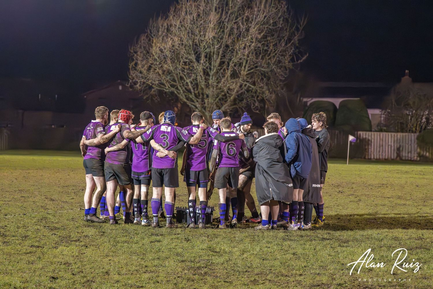 Rugby teams huddle together on the field after a match as the sun sets.