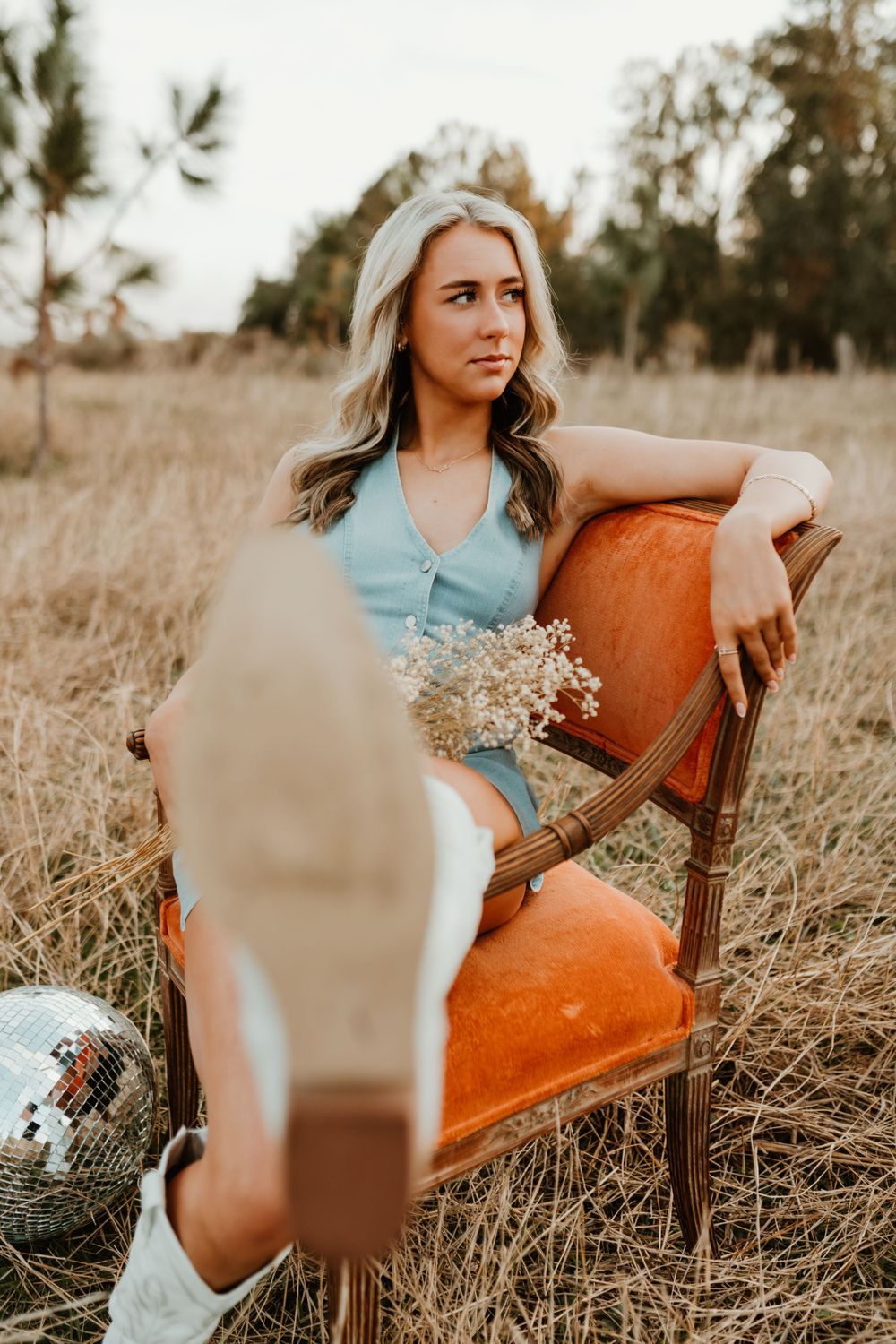 Woman in blue dress poses on vintage orange chair in field with cowboy boots.