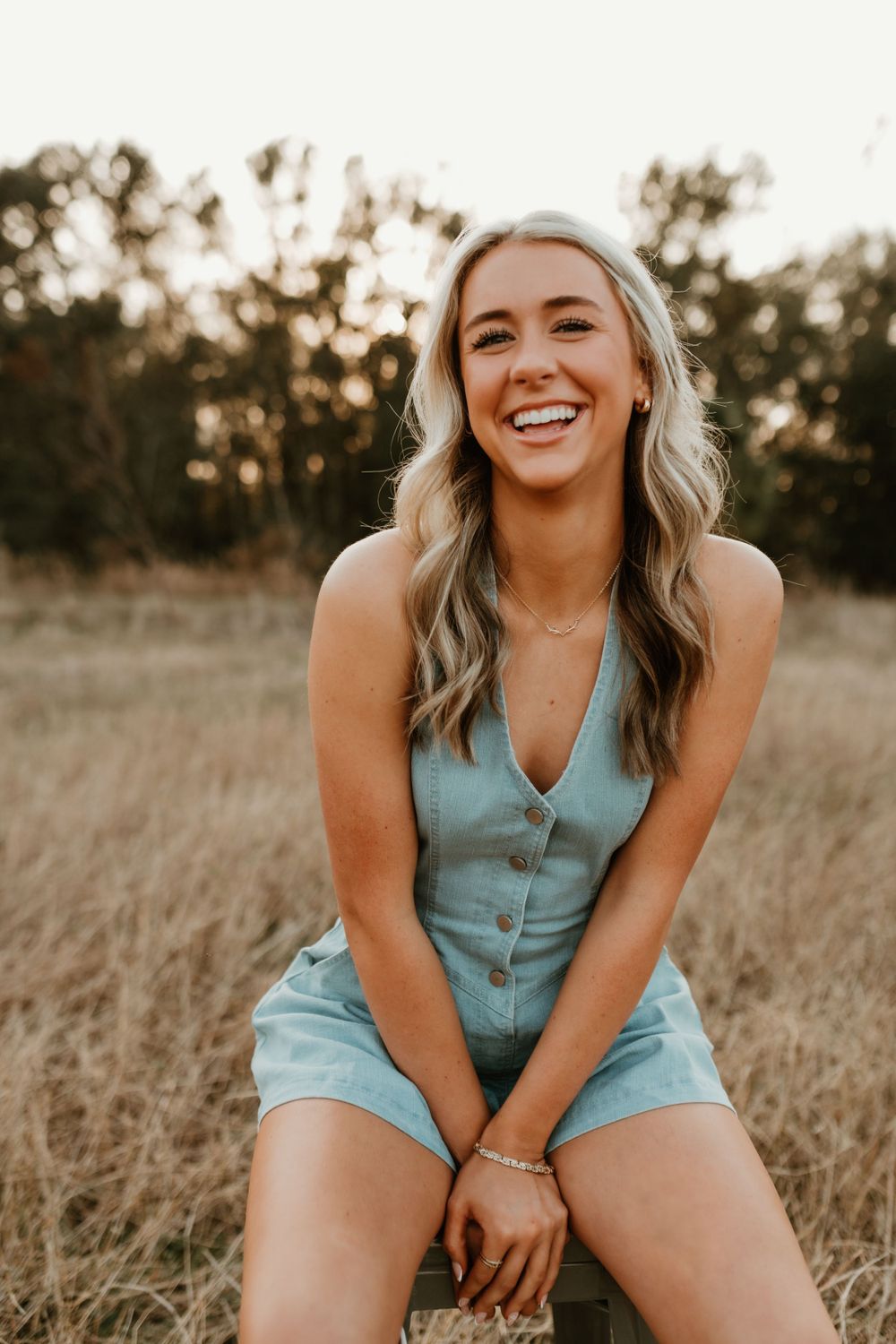 Person wearing blue romper laughs while sitting in tall grass during golden hour.
