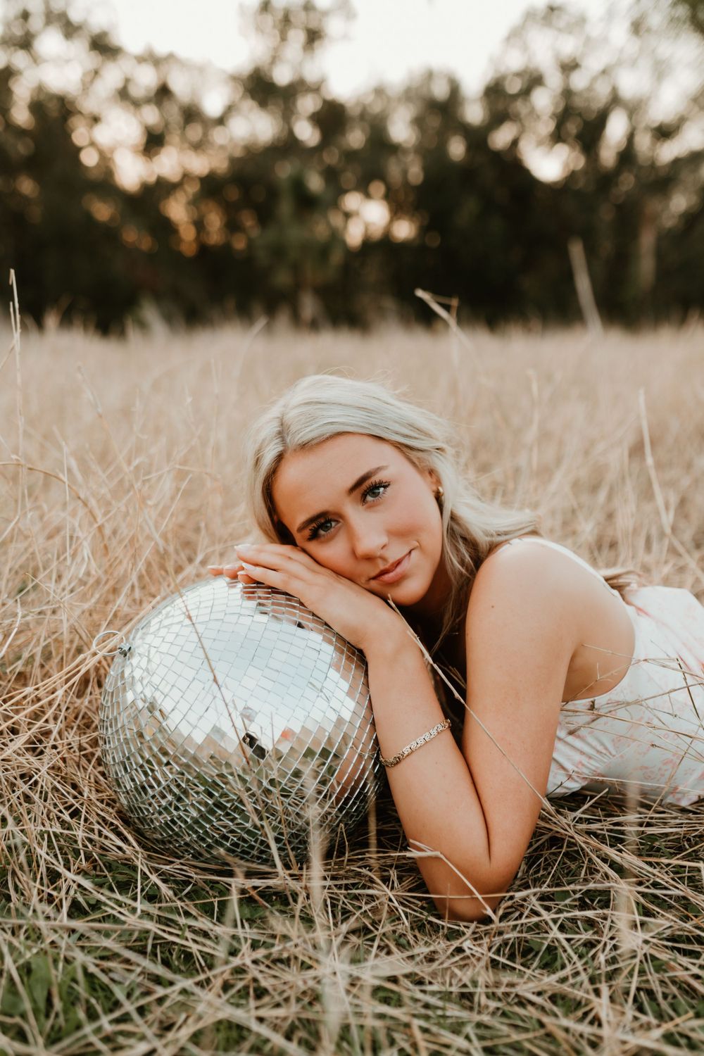 Person poses with disco ball in dried grass field during sunset photoshoot.