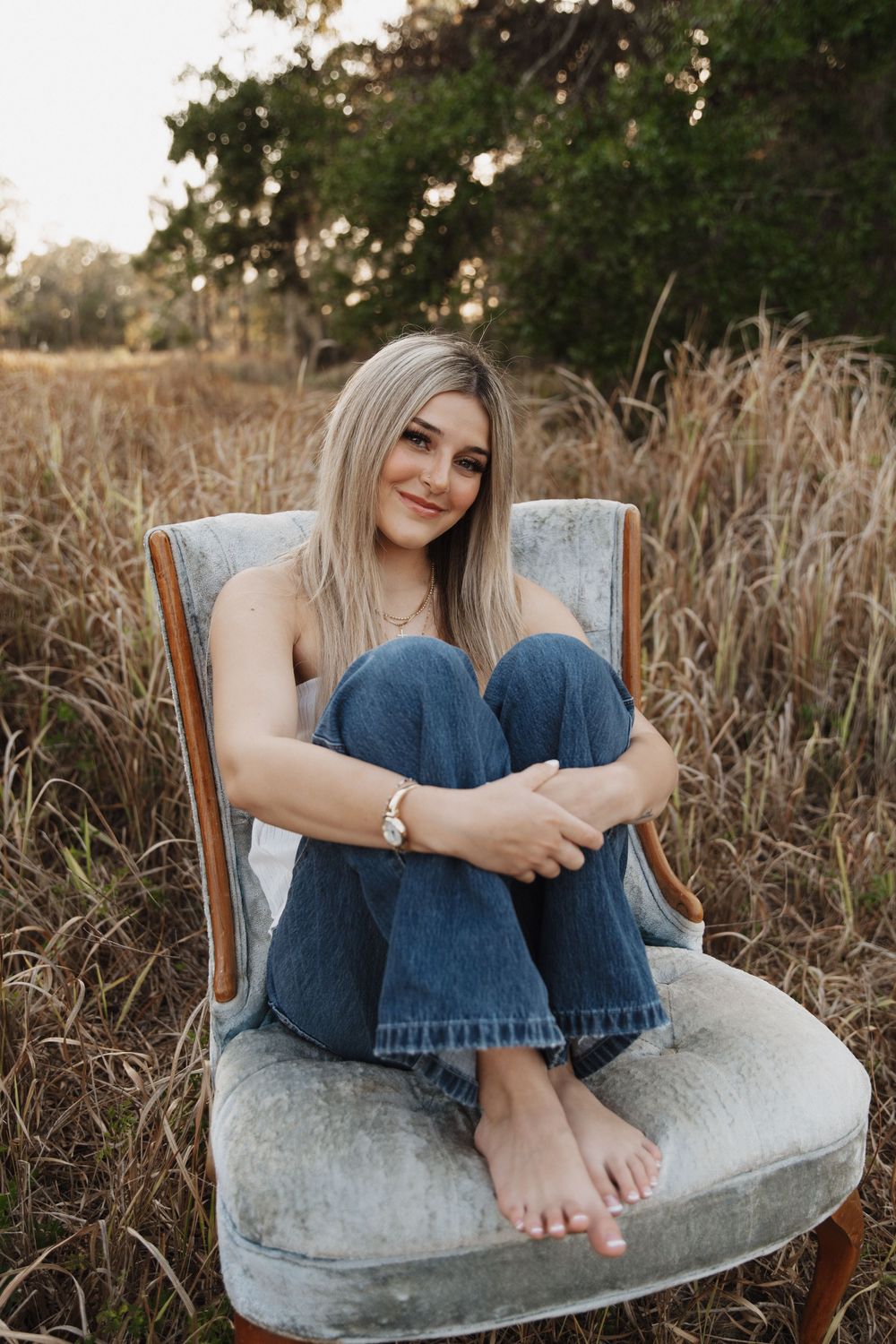 Carefree graduate relaxes barefoot on vintage chair outdoors among tall grass during sunset photoshoot.