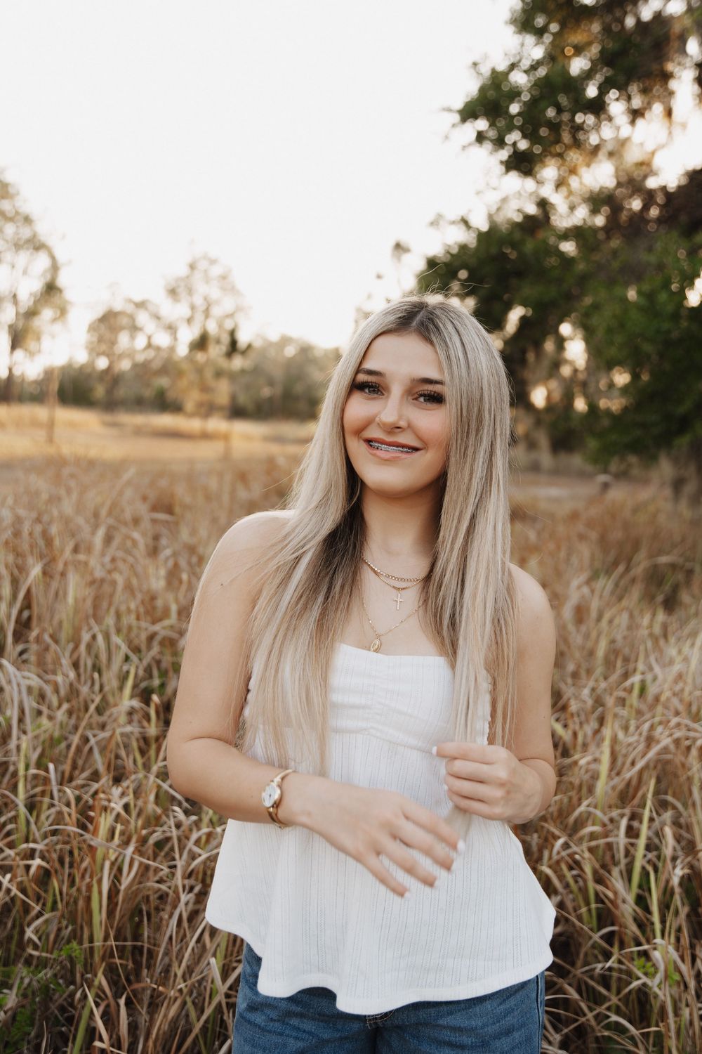 A woman in a white top poses in a golden field during sunset.
