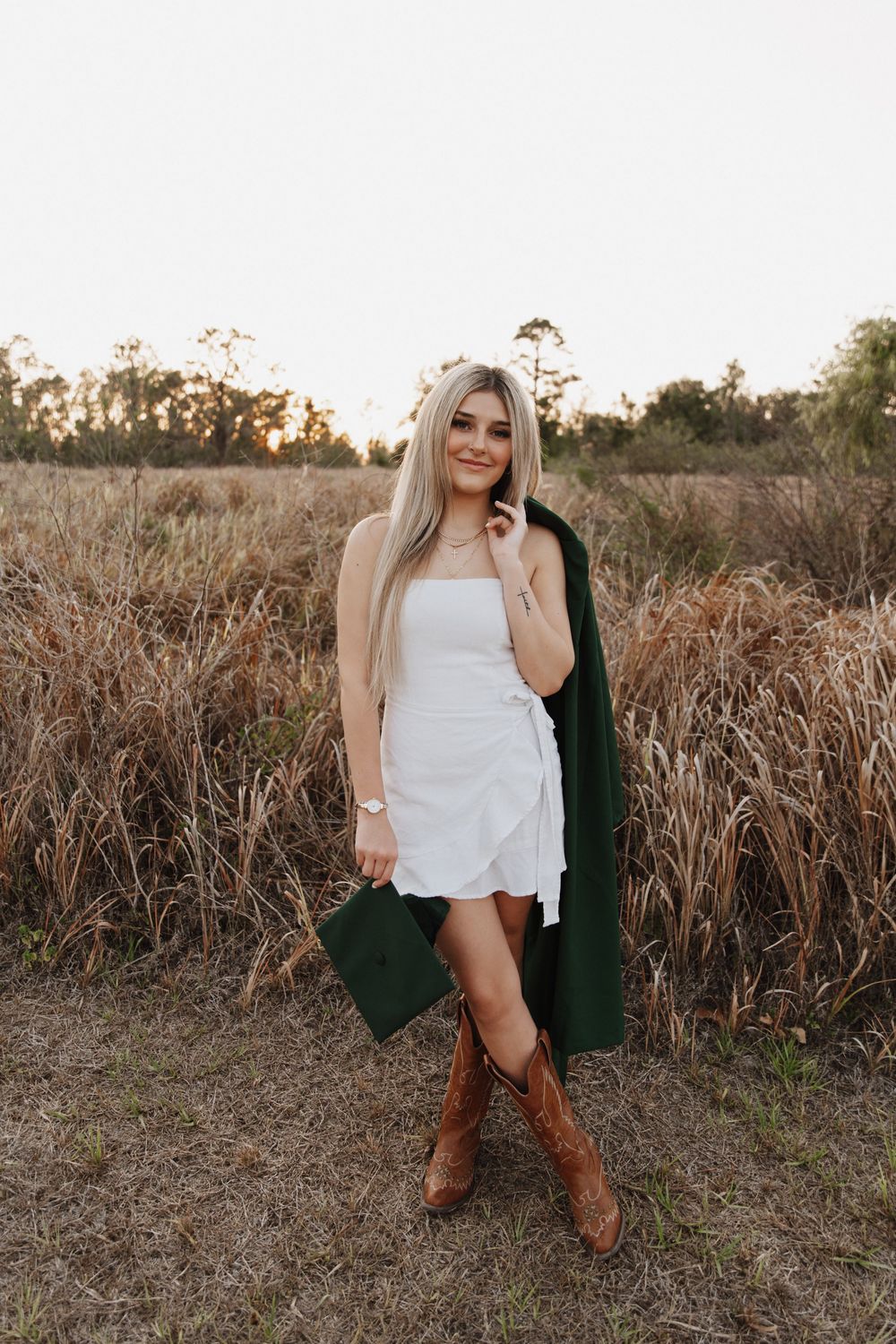 A person in a white dress and dark cardigan poses amid tall dried grass during golden hour.