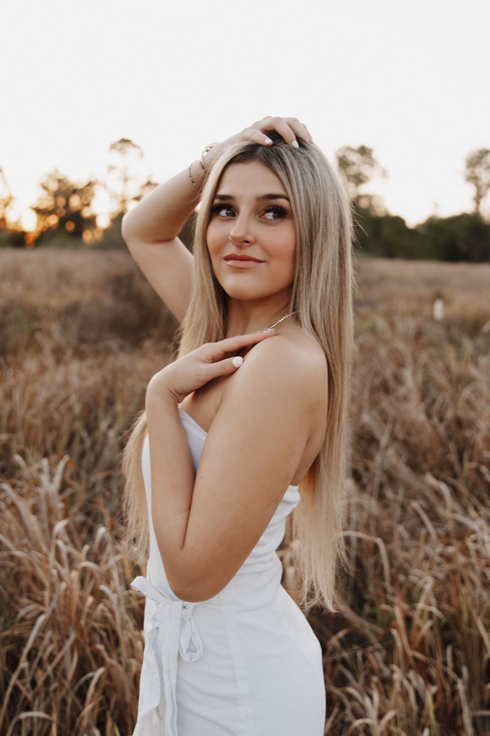 A blonde woman in a white sundress poses gracefully in a golden wheat field during sunset.