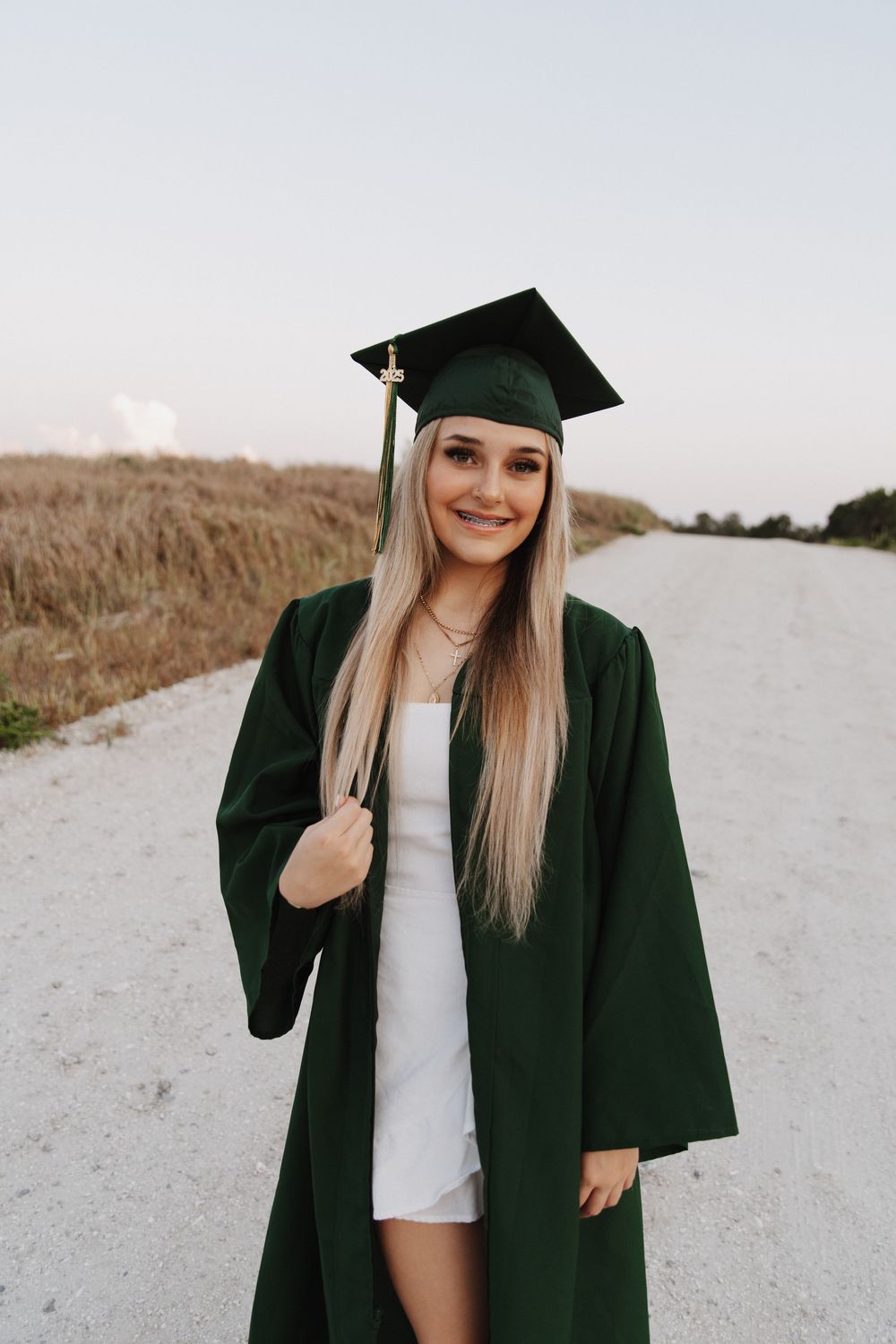 Graduate in dark green cap and gown poses on dirt path outdoors during golden hour portrait session.