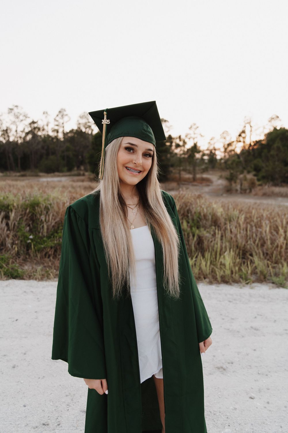 Graduate in green cap and gown poses outdoors during golden hour on sandy beach with tall grass.