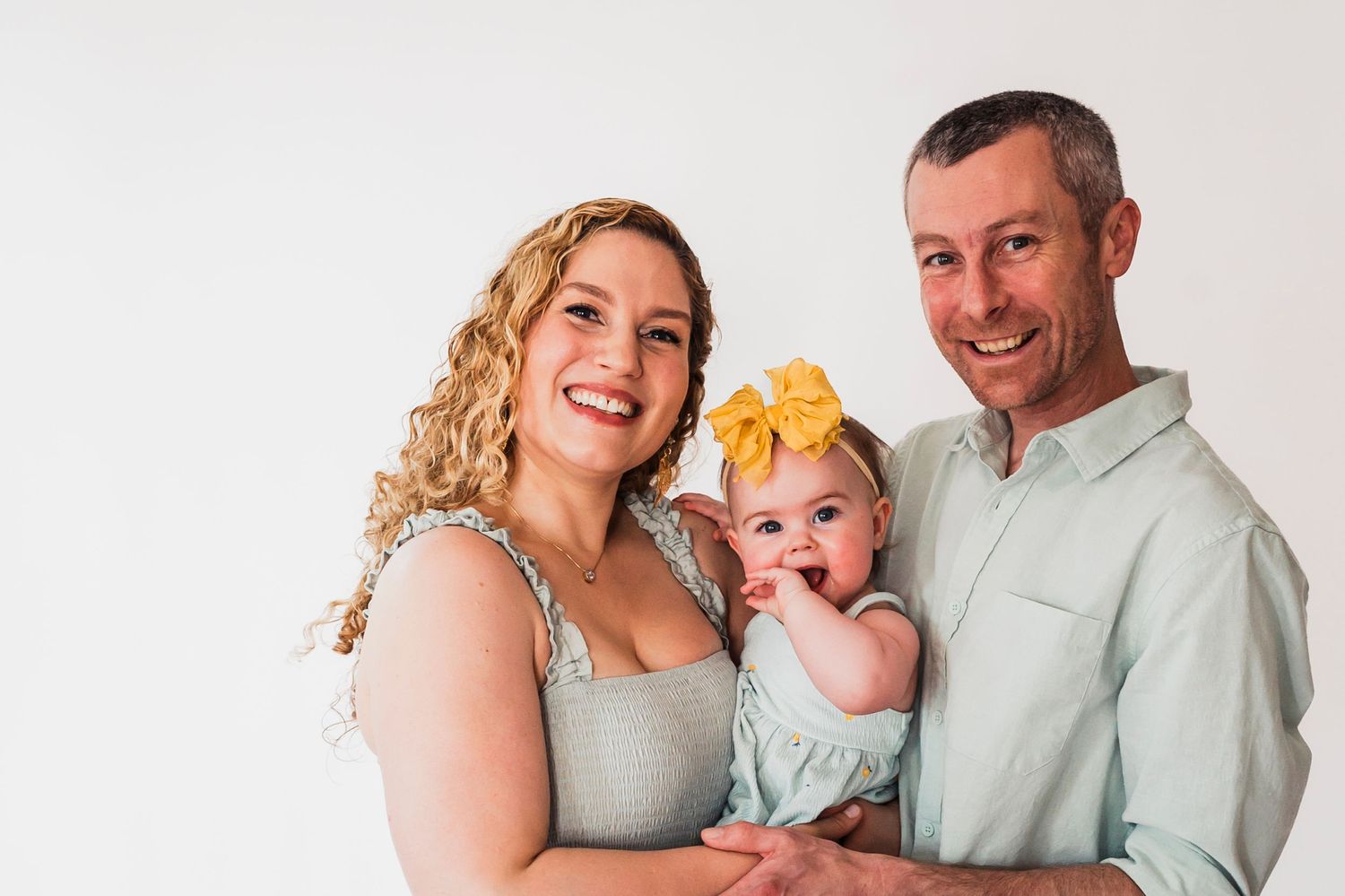 A family portrait against a white background features parents in light colored clothing holding their baby with a yellow headband.