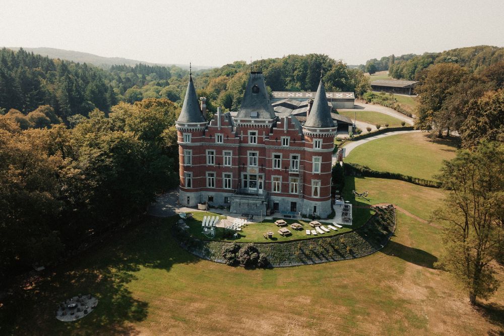 Aerial view of a Gothic-style red brick castle with twin spires surrounded by rolling green lawns and woodland.