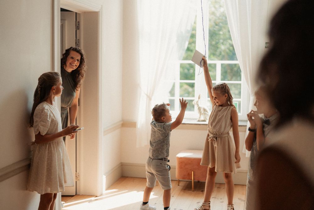 Sunlit room with children playing near curtained windows while adults watch from doorway.