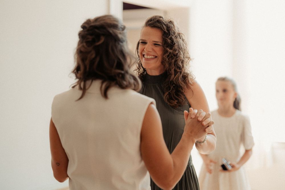 Two people dance together in a bright room while another watches from a distance in the background.