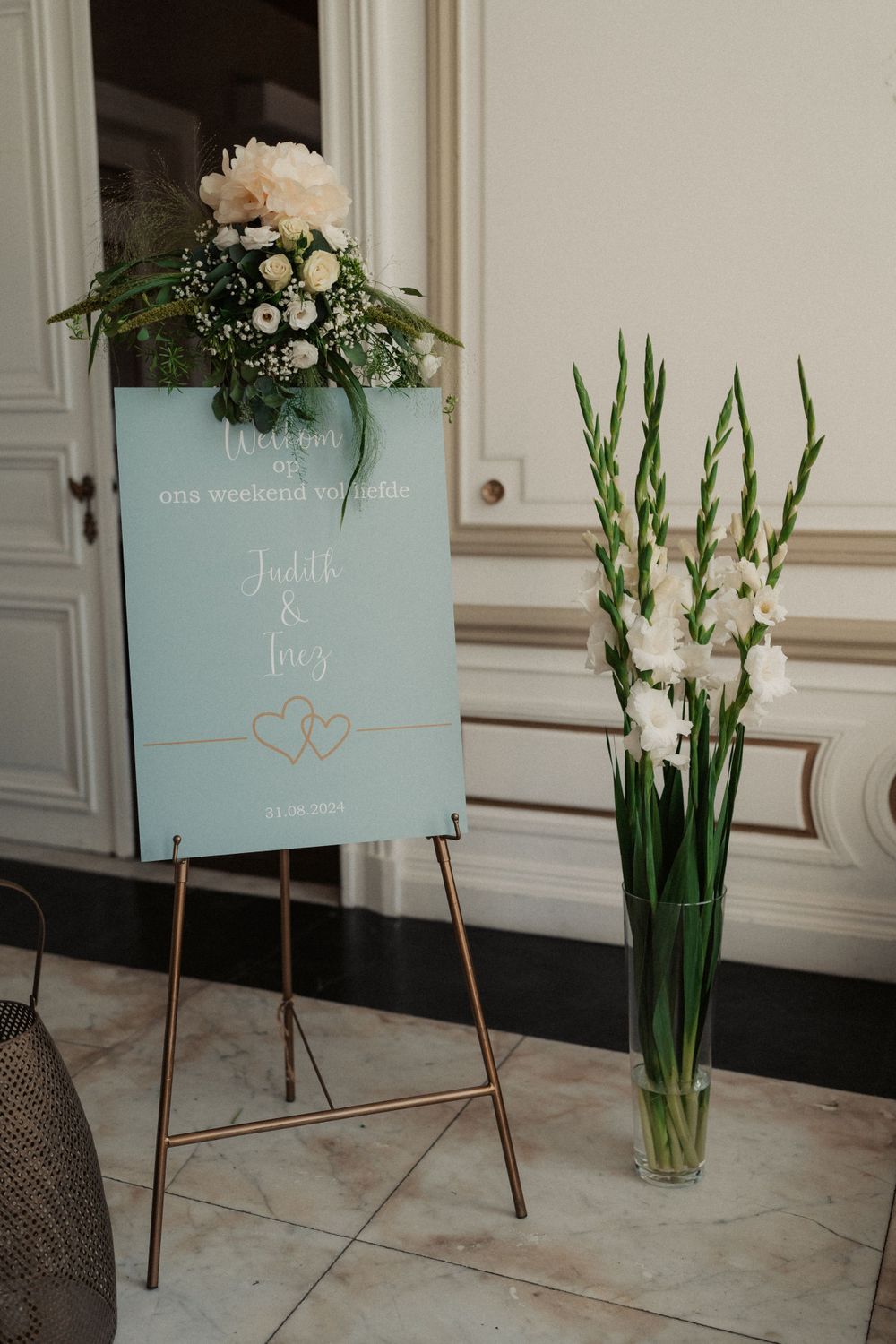 Elegant mint green wedding welcome sign with floral arrangement and gold easel stand next to white gladiolus in a glass vase.