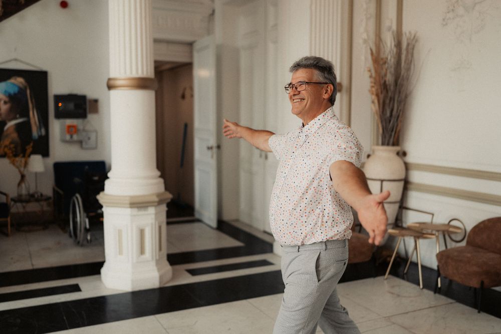 A person in glasses and a polka dot shirt strikes a happy, playful pose in an elegant hallway with white columns.